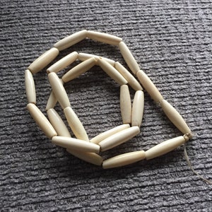 Puede incluir: Una hebra de cuentas cilíndricas de color blanco hueso hechas de hueso o marfil. Las cuentas están enhebradas con un cordón blanco delgado.