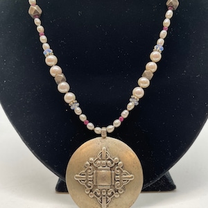 May include: A silver necklace with a large, round pendant featuring a geometric design. The necklace is made of white pearls, small pink and blue beads.