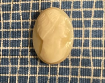 May include: Oval-shaped cameo in cream color, featuring a profile of a woman's face. The cameo is set against a blue and white checkered background. The carving details are visible, showcasing the woman's hair and facial features.