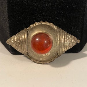 May include: A silver-toned metal brooch with a red glass cabochon in the center. The brooch has a decorative design around the edges.