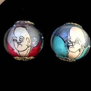 May include: Two round glass beads with gold trim, each containing a painted image of a person with a white face and black hair. One bead has a red background and the other has a blue background.