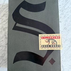 May include: A rectangular, dark gray book with a large black letter "S" on the cover. A sticker with the text "J.J. ABRAMS DOUG DORST" and a monkey illustration is affixed to the front. The book has a diamond shape at the bottom.