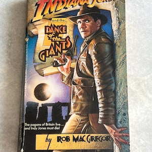 May include: A paperback book cover featuring Indiana Jones, the character from the movie franchise, holding a gun and standing in front of a stone structure. The title of the book is "Indiana Jones and the Dance of the Giants". The book cover is illustrated with a dark blue background and a bright yellow sky.