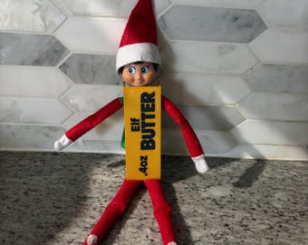 Pode incluir: Uma boneca elfo articulada com gorro vermelho e roupa vermelha, usando uma placa retangular amarela no peito com a inscrição Elf BUTTER .4oz em preto. A boneca tem olhos azuis e punhos brancos nas mangas e nas pernas.