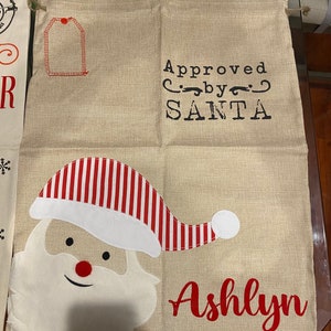 Pode incluir: Uma meia de Natal de estopa bege com um chap&eacute;u de Papai Noel listrado de vermelho e branco, uma barba branca e o texto "Approved by SANTA" e "Ashlyn".