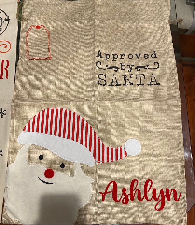 Pode incluir: Uma meia de Natal de estopa bege com um chap&eacute;u de Papai Noel listrado de vermelho e branco, uma barba branca e o texto "Approved by SANTA" e "Ashlyn".