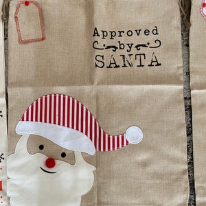 Pode incluir: Uma sacola de presente de juta bege com um chap&eacute;u de Papai Noel listrado vermelho e branco e uma barba branca. A sacola tem uma etiqueta e o texto "Aprovado por SANTA".