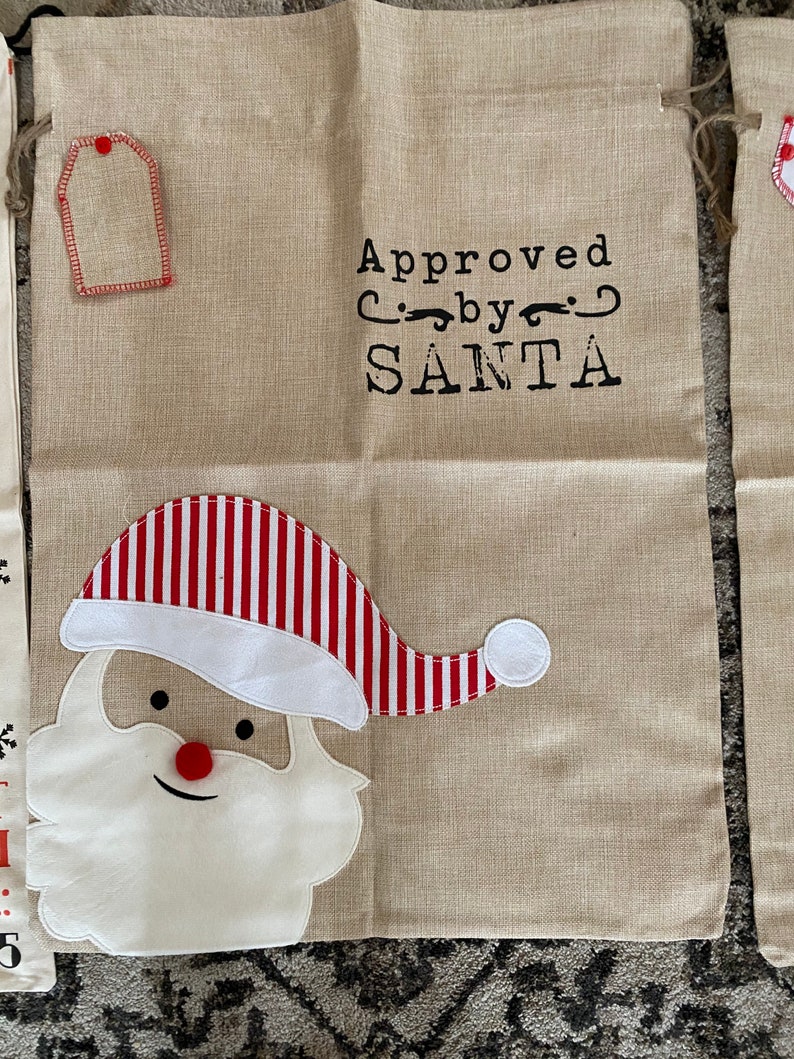 Pode incluir: Uma sacola de presente de juta bege com um chap&eacute;u de Papai Noel listrado vermelho e branco e uma barba branca. A sacola tem uma etiqueta e o texto "Aprovado por SANTA".