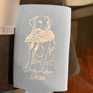 May include: A light blue foam can cooler featuring a white line art illustration of a dog holding a duck in its mouth. Below the illustration, the text reads "Drinks on Aspen" in a cursive script, followed by the date "5.9.26" in a bold font.