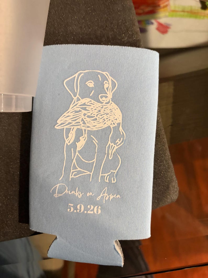 May include: A light blue foam can cooler featuring a white line art illustration of a dog holding a duck in its mouth. Below the illustration, the text reads "Drinks on Aspen" in a cursive script, followed by the date "5.9.26" in a bold font.