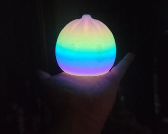 Pode incluir: Uma pequena lâmpada de silicone redonda e brilhante em forma de pão cozido no vapor ou bolinho. A superfície emite uma luz suave e multicolorida, que transita do amarelo no topo para o verde, azul e roxo na base, criando um efeito de gradiente.