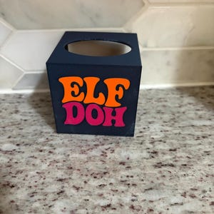 May include: A dark blue square container with a circular opening on the top surface. The front face features the text "ELF DOH" in a bold, stylized font, with "ELF" printed in bright orange and "DOH" printed in a vibrant pink color.