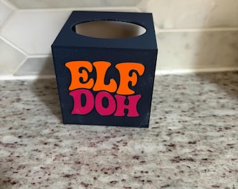 Pode incluir: Um recipiente quadrado azul-escuro com uma abertura circular na parte superior. A face frontal apresenta o texto "ELF DOH" numa fonte negrito e estilizada, com "ELF" impresso em laranja brilhante e "DOH" em cor-de-rosa vibrante.