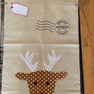 Pode incluir: Uma bolsa de presente de juta bege com uma aplica&ccedil;&atilde;o de rosto de rena. A rena tem pele marrom com bolinhas brancas e chifres vermelhos com um padr&atilde;o xadrez. A bolsa tem uma etiqueta branca com uma borda vermelha e um carimbo preto que diz "Reindeer Mail North Pole Express".