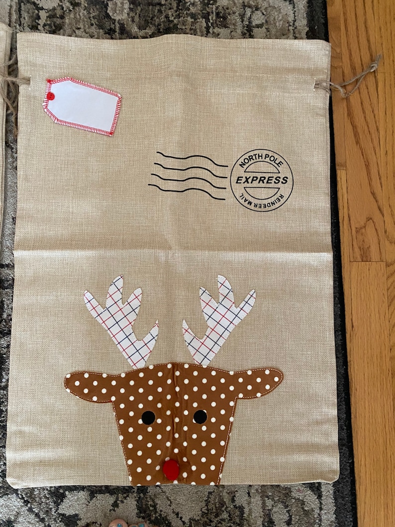 Pode incluir: Uma bolsa de presente de juta bege com uma aplica&ccedil;&atilde;o de rosto de rena. A rena tem pele marrom com bolinhas brancas e chifres vermelhos com um padr&atilde;o xadrez. A bolsa tem uma etiqueta branca com uma borda vermelha e um carimbo preto que diz "Reindeer Mail North Pole Express".