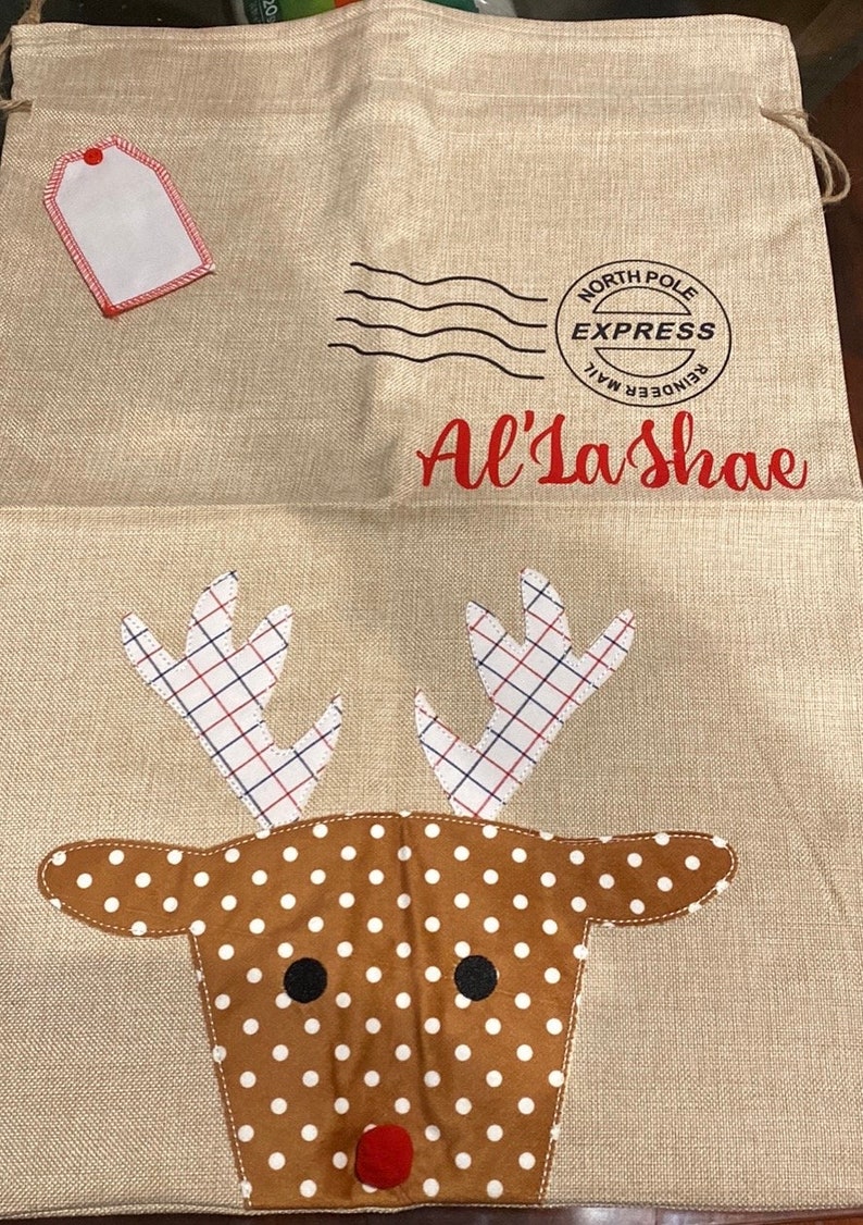 Pode incluir: Uma sacola de presente de Natal de estopa bege com um rosto de rena vermelho e chifres. A sacola tem uma etiqueta vermelha e um carimbo preto que diz "North Pole Express Reindeer Mail". A sacola &eacute; personalizada com o nome "Al'Lashae".