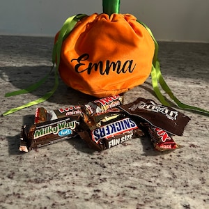 Pu&ograve; includere: Borsa di stoffa arancione a forma di zucca con nastri verdi e il nome "Emma" ricamato sul davanti. La borsa &egrave; piena di barrette di cioccolato assortite, tra cui Milky Way, Snickers e Twix.