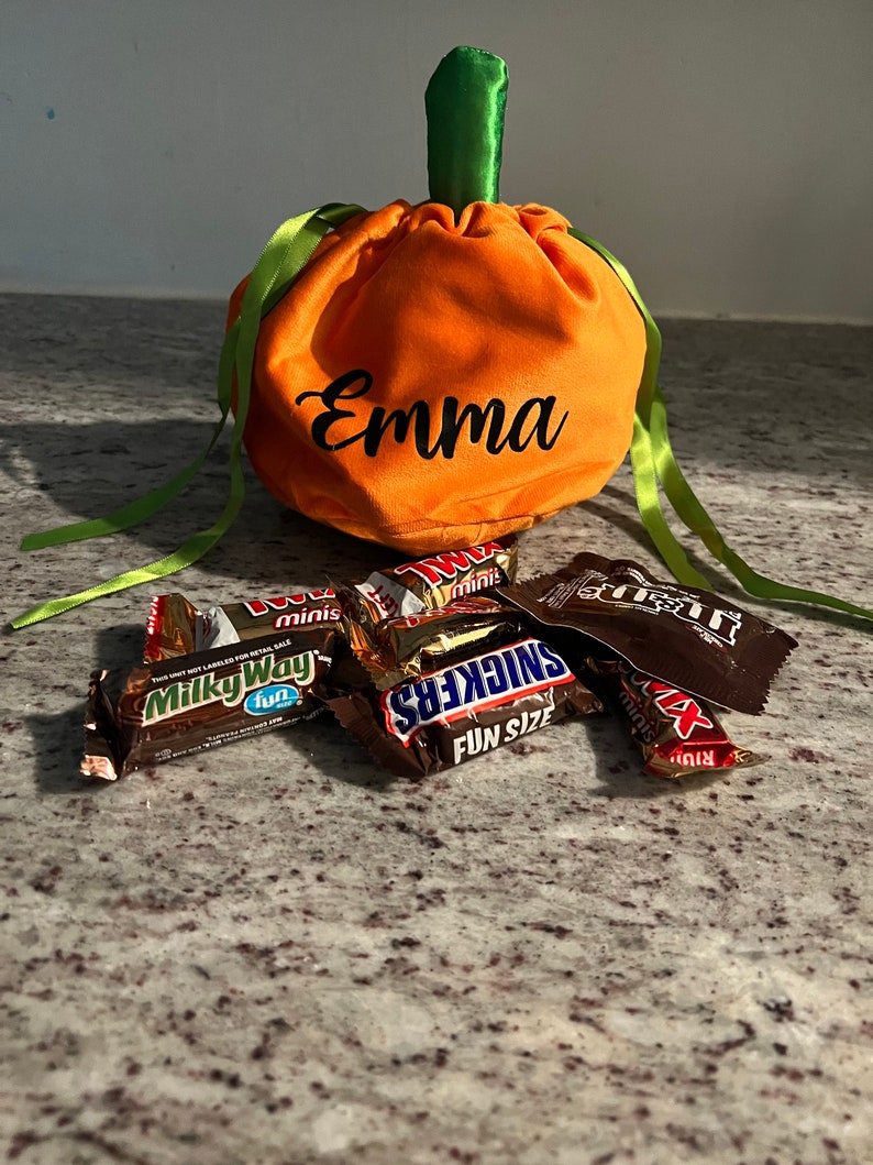 Pu&ograve; includere: Borsa di stoffa arancione a forma di zucca con nastri verdi e il nome "Emma" ricamato sul davanti. La borsa &egrave; piena di barrette di cioccolato assortite, tra cui Milky Way, Snickers e Twix.