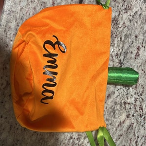 Pu&ograve; includere: Borsa di stoffa arancione a forma di zucca con lacci di nastro verde. La borsa ha la scritta "Autunno" in nero.