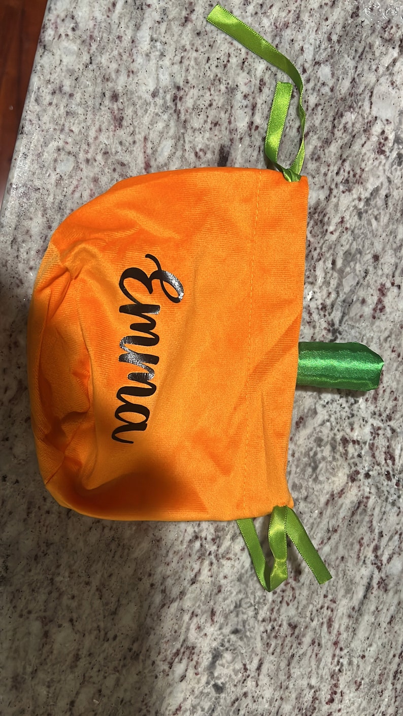 Pu&ograve; includere: Borsa di stoffa arancione a forma di zucca con lacci di nastro verde. La borsa ha la scritta "Autunno" in nero.