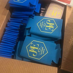May include: A collection of blue foam can coolers packed in a cardboard box. Each cooler features a gold hexagonal emblem with the initials J&E, a heart icon, and the text THE HAGERS 10.04.25. One tan cooler with the same gold design is also visible.