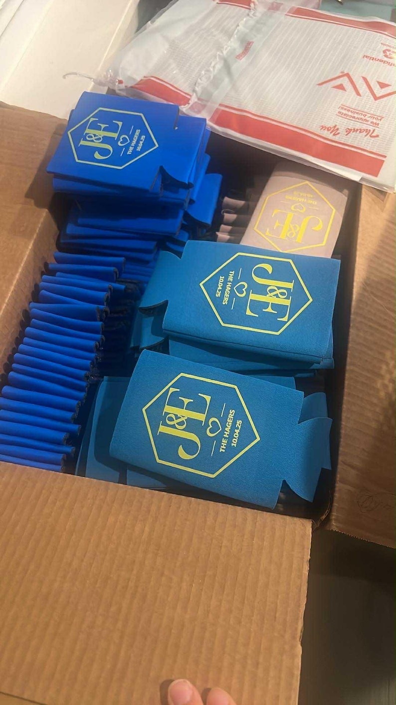 May include: A collection of blue foam can coolers packed in a cardboard box. Each cooler features a gold hexagonal emblem with the initials J&E, a heart icon, and the text THE HAGERS 10.04.25. One tan cooler with the same gold design is also visible.