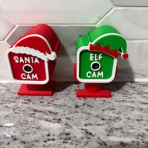 May include: Two decorative camera ornaments with stands. The red one features a white Santa hat and the text SANTA CAM. The green one features a red and green elf hat and the text ELF CAM. Both have a central black circular lens detail.