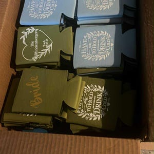 May include: A collection of foam can coolers in light blue and olive green colors, stacked inside a cardboard box. The coolers feature white printed text including phrases like To Have and To Hold and To Keep Your Drink Cold, along with custom names.