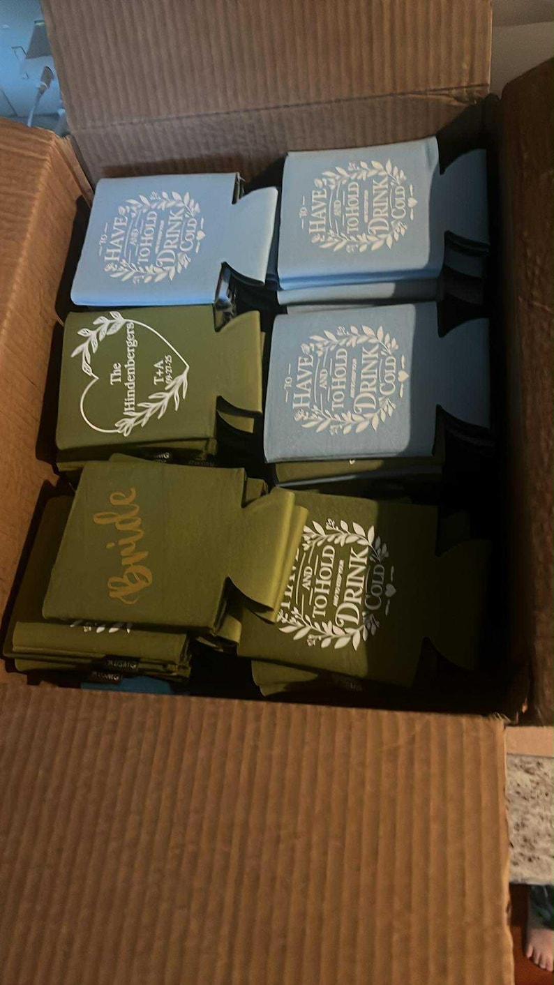 May include: A collection of foam can coolers in light blue and olive green colors, stacked inside a cardboard box. The coolers feature white printed text including phrases like To Have and To Hold and To Keep Your Drink Cold, along with custom names.