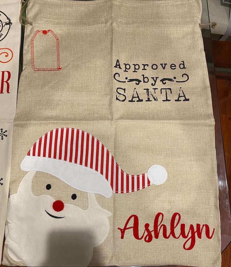 Pode incluir: Uma meia de Natal de estopa bege com um chap&eacute;u de Papai Noel listrado de vermelho e branco, uma barba branca e o texto "Approved by SANTA" e "Ashlyn".