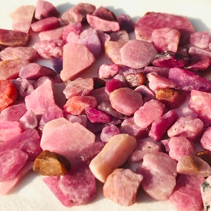 Puede incluir: Un conjunto de piedras preciosas en bruto, sin tallar, en tonos rosas y rojos. Las piedras varían en tamaño y forma, con una mezcla de tonos claros y oscuros. Algunas piedras son translúcidas, otras opacas.