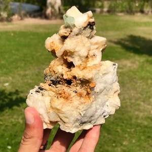 May include: A natural mineral specimen featuring a cluster of off-white, tan, and black rock formations. A small, light green crystal is visible at the top. The rough texture and varied colors create a unique display.