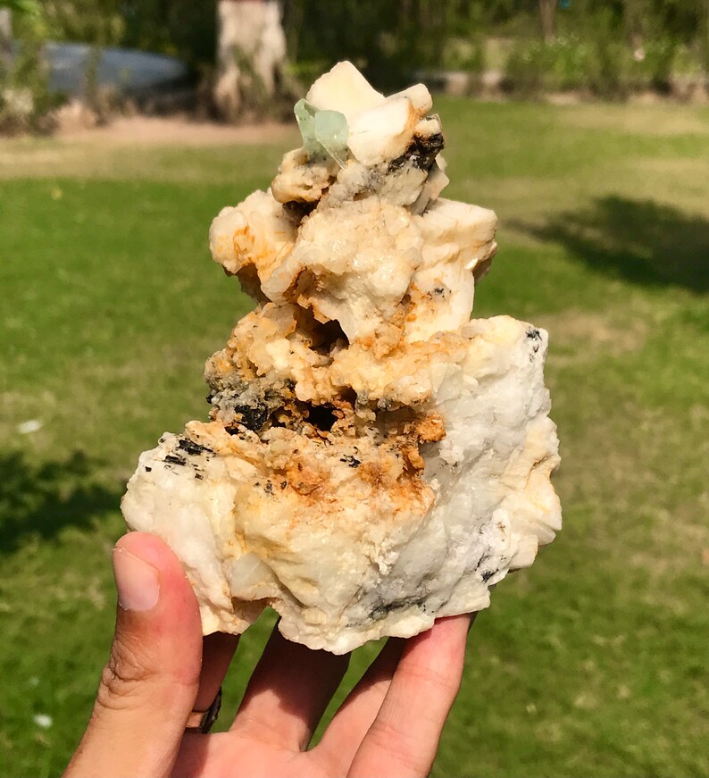May include: A natural mineral specimen featuring a cluster of off-white, tan, and black rock formations. A small, light green crystal is visible at the top. The rough texture and varied colors create a unique display.