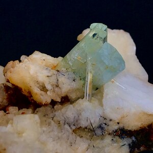 May include: A close-up of a mineral specimen featuring a cluster of pale blue-green aquamarine crystals. The crystals are set against a backdrop of white and beige matrix, with dark inclusions.