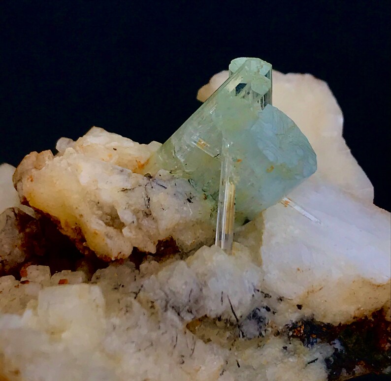 May include: A close-up of a mineral specimen featuring a cluster of pale blue-green aquamarine crystals. The crystals are set against a backdrop of white and beige matrix, with dark inclusions.