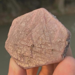 Könnte beinhalten: Ein hexagonales Rohkristall-Exemplar in einem staubigen Rosaton. Die Oberfläche zeigt ein komplexes Netzwerk aus sich kreuzenden linearen Rillen und dreieckigen Mustern, was ein texturiertes, mattes Erscheinungsbild auf der flachen Seite erzeugt.