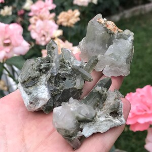 May include: A collection of raw quartz crystals in shades of clear, gray, and green. The crystals are clustered together, showcasing their natural, rough texture. The background features soft pink and beige flowers.