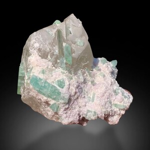 Peut inclure: Spécimen minéral naturel avec des cristaux de tourmaline vert clair incrustés dans une matrice de quartz et d'un minéral blanc-rosâtre pâle. Les cristaux sont allongés et prismatiques, d'aspect translucide. Fond noir.