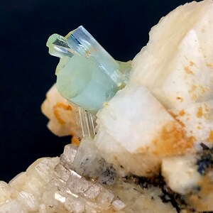 May include: Close-up of a mineral specimen featuring a pale blue aquamarine crystal formation. The aquamarine crystals are translucent and sit atop a matrix of white and beige minerals, with a dark background.