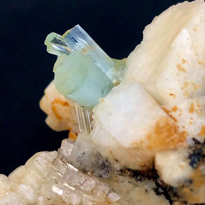 May include: Close-up of a mineral specimen featuring a pale blue aquamarine crystal formation. The aquamarine crystals are translucent and sit atop a matrix of white and beige minerals, with a dark background.