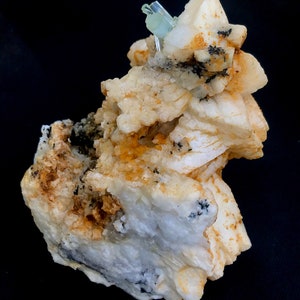 May include: A close-up of a mineral specimen featuring a cluster of off-white, beige, and tan crystals. Small, light blue crystals are at the top. Black flecks are scattered throughout the specimen. The background is black.