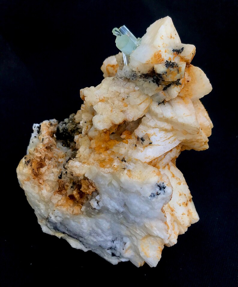 May include: A close-up of a mineral specimen featuring a cluster of off-white, beige, and tan crystals. Small, light blue crystals are at the top. Black flecks are scattered throughout the specimen. The background is black.