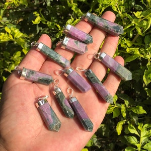 May include: A collection of gemstone pendants in various sizes, held in a hand. The pendants are a mix of pink and green hues, with silver-colored metal caps and loops for hanging. The background is a blurred view of green foliage.