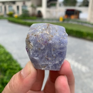 May include: A rough, uncut gemstone with a light purple hue. The stone has a textured surface with small flecks of darker color. The gemstone is held in a person's hand, with a blurred outdoor background.
