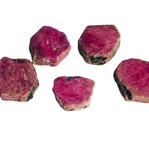 Puede incluir: Cinco piedras preciosas de rubí en bruto, de un color rojo rosado intenso. Las piedras tienen una superficie texturizada y formas irregulares, algunas muestran inclusiones de color verde oscuro. Las piedras se muestran sobre un fondo blanco.