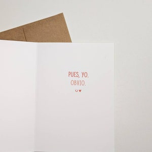 May include: A white card with the text "Pues, yo. Obvio." in pink. The card is folded in half and has a brown envelope behind it.