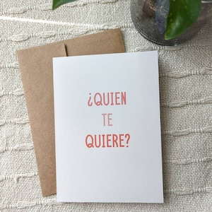 May include: A white card with the text "&iquest;QUIEN TE QUIERE?" in pink lettering. The card is sitting on a beige textured surface.