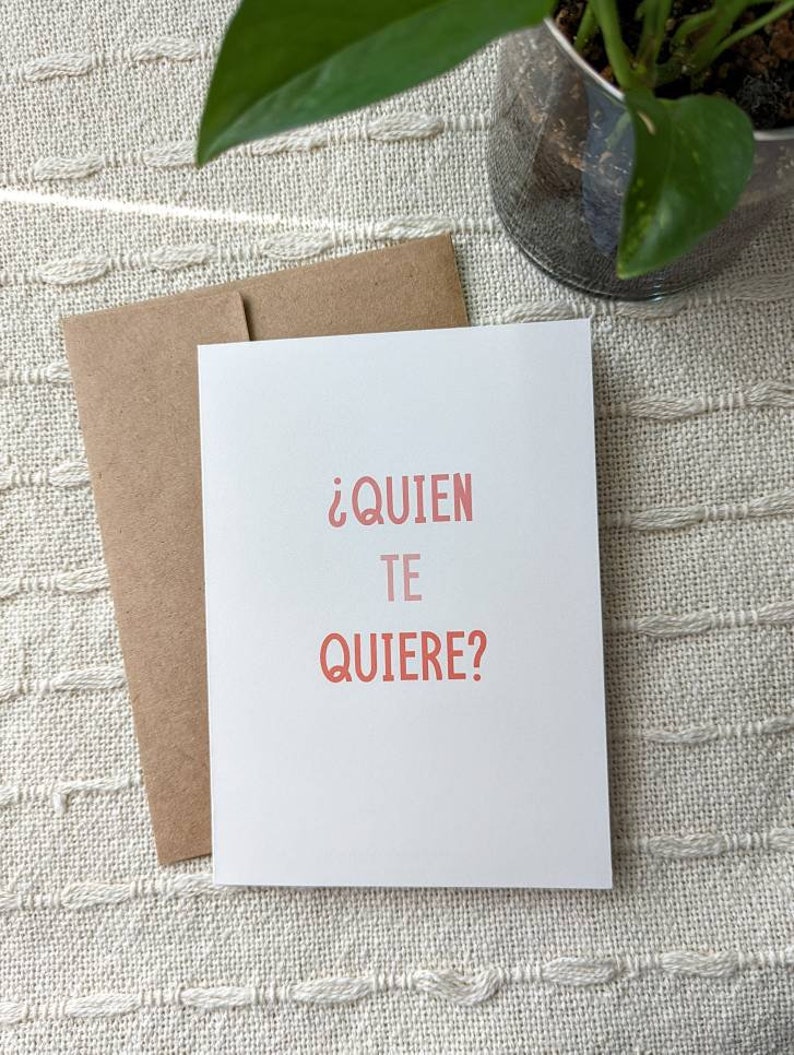 May include: A white card with the text "&iquest;QUIEN TE QUIERE?" in pink lettering. The card is sitting on a beige textured surface.