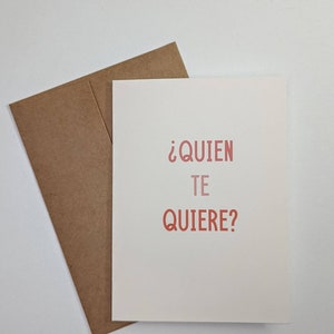 May include: A white card with a brown envelope. The card has the text "&iquest;QUIEN TE QUIERE?" in red.