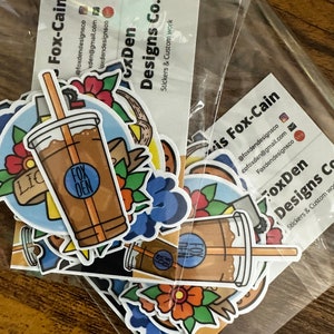 May include: A set of stickers featuring a cartoon illustration of a coffee cup with a straw, a flower, and the text "Fox Den Designs Co.". The stickers are packaged in a clear plastic bag.